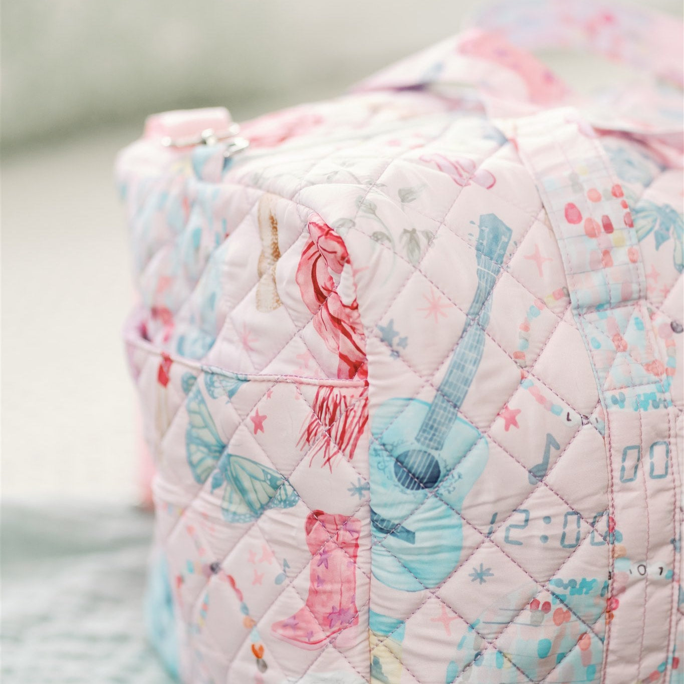 Light pink quilted duffle with 2 handles, a strap, and a pattern featuring original watercolor art of Taylor Swift icons.