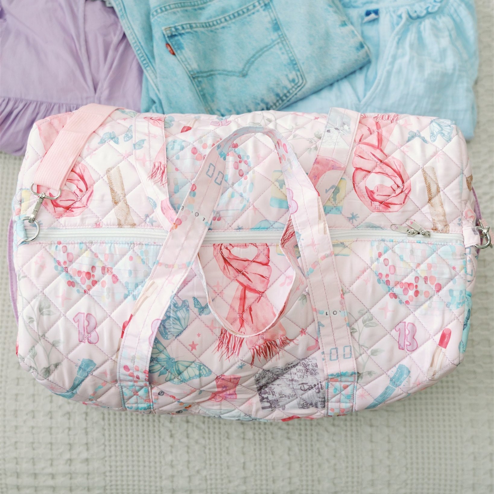 Light pink quilted duffle with 2 handles, a strap, and a pattern featuring original watercolor art of Taylor Swift icons.