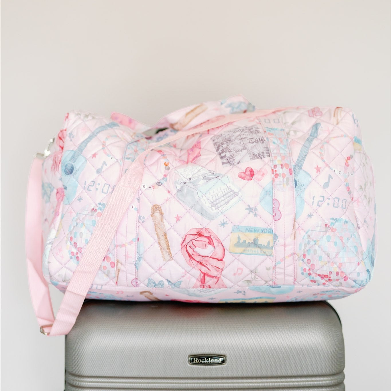 Light pink quilted duffle with 2 handles, a strap, and a pattern featuring original watercolor art of Taylor Swift icons.