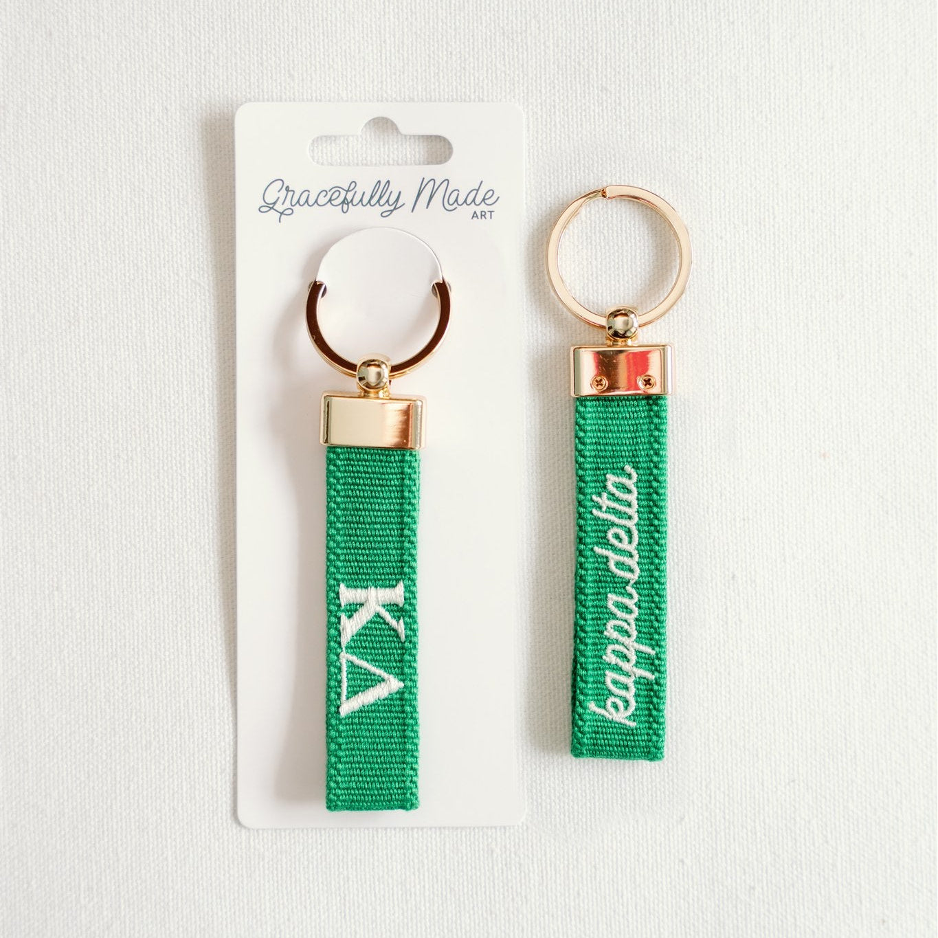 Front and back of a green keychain with Kappa Delta embroidered in white in Gracefully Made Art branded packaging.