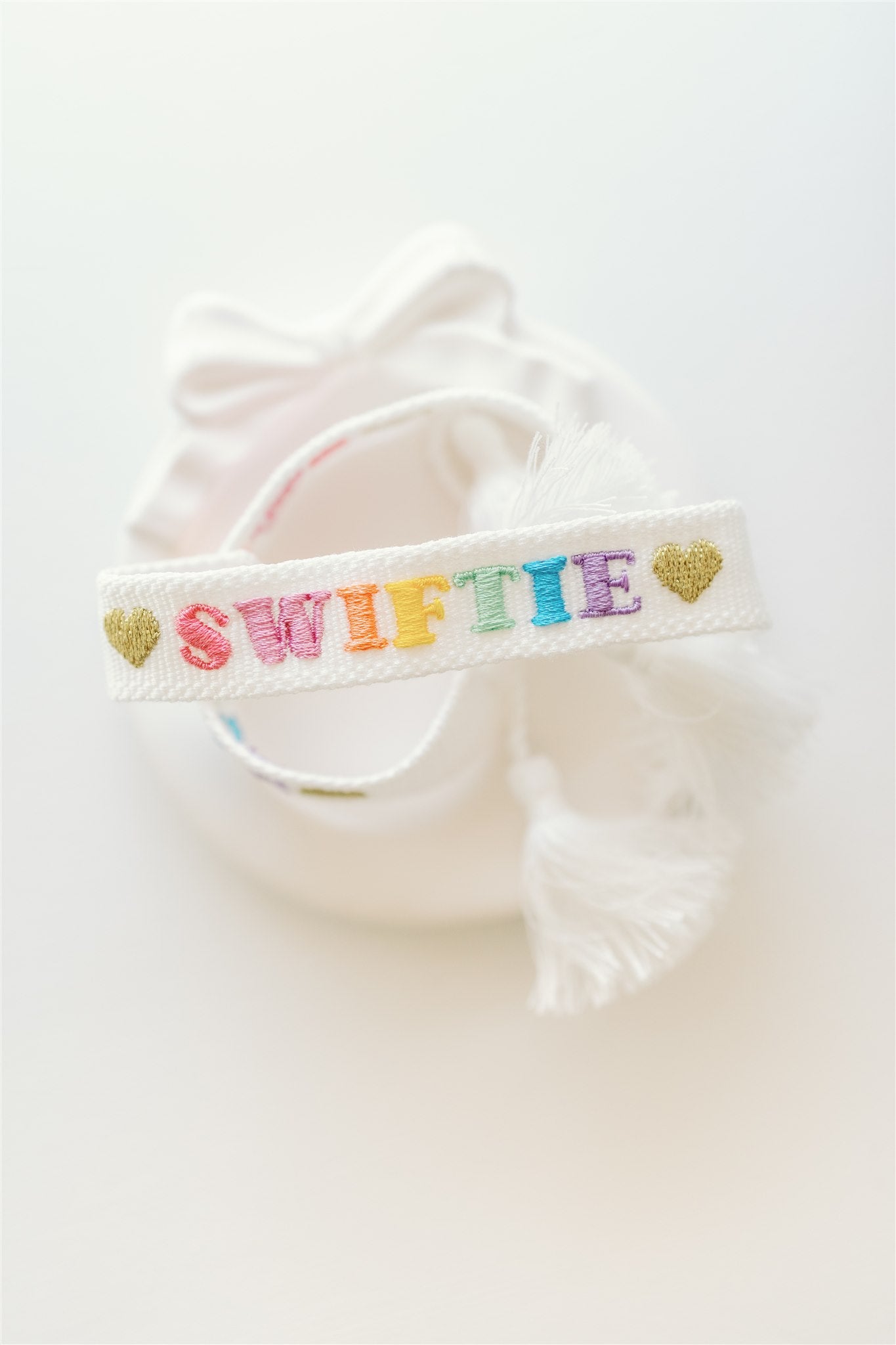 White bracelet with "Swiftie" embroidered on it in different colors and finished with gold hearts.