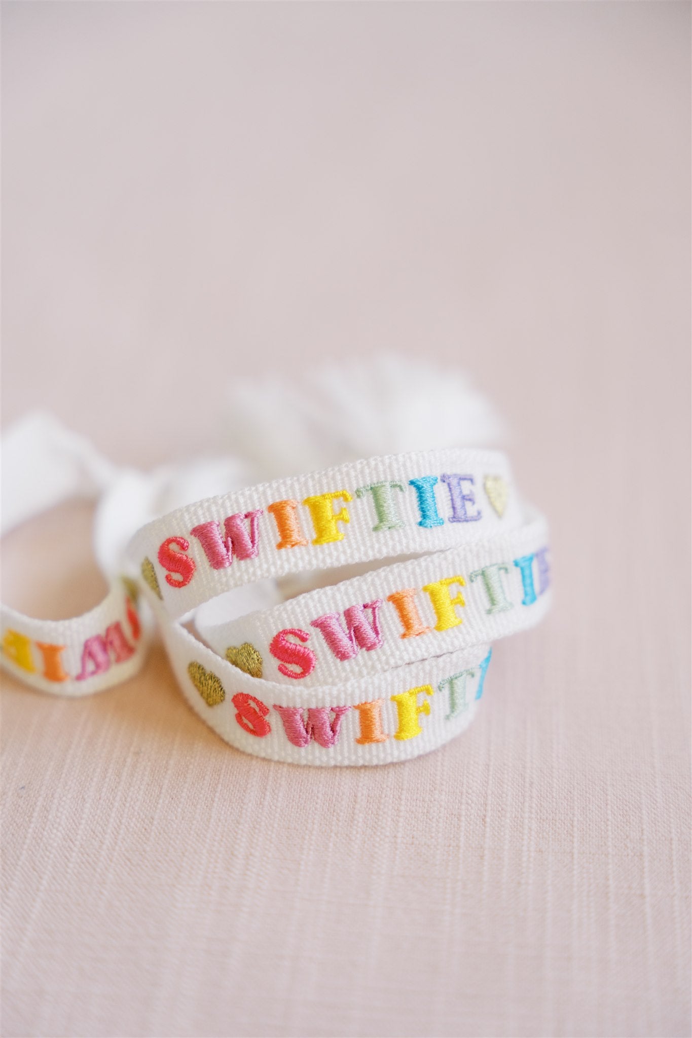 White bracelet with "Swiftie" embroidered on it in different colors and finished with gold hearts.