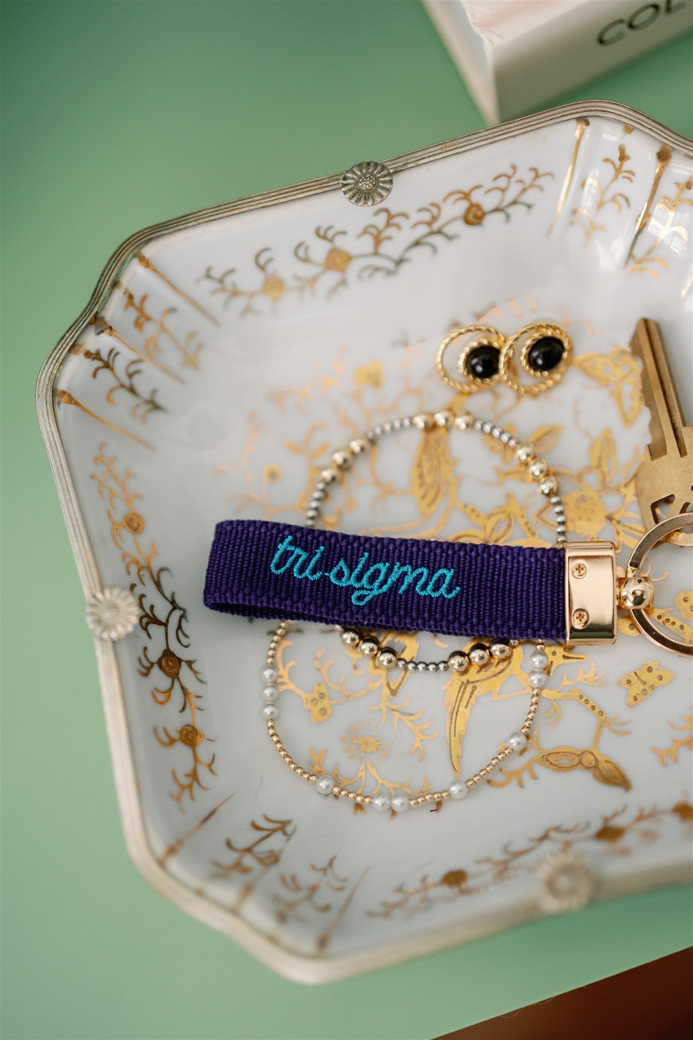 Decorative dish with jewelry including a key, bracelet, and 'tri sigma' keychain on a green background.