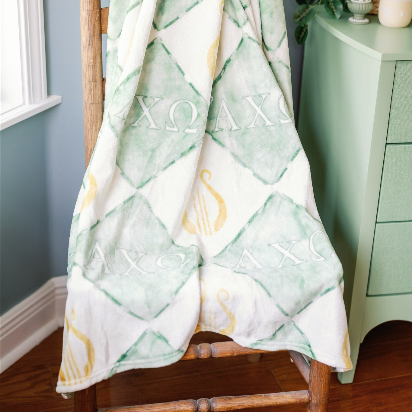 Green and white Alpha Chi Omega patterned blanket draped over a wooden chair against a gray wall.