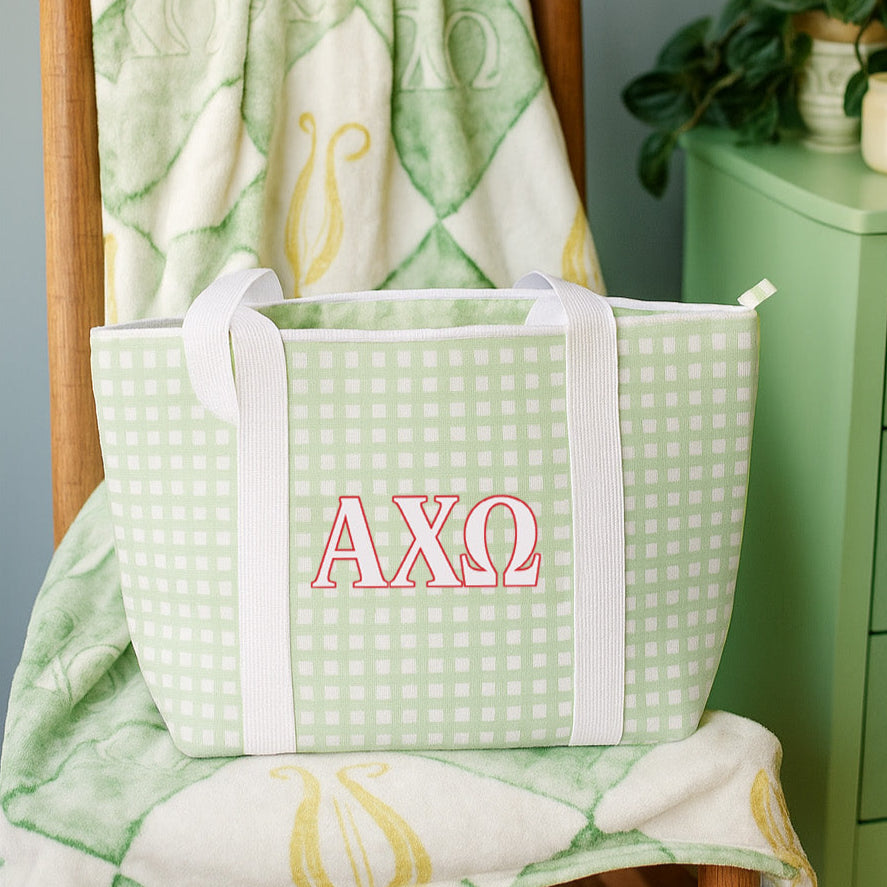 Green bag with Alpha Chi Omega Greek letters on a wooden chair against a patterned blanket.