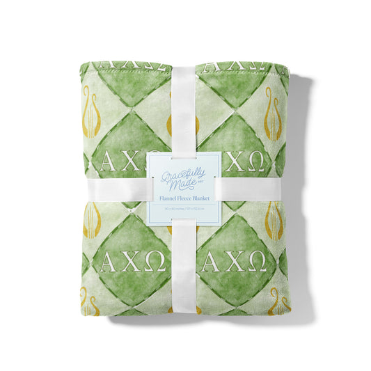 Folded green and white patterned blanket with 'AXOmega' symbols, branded 'Handfuls Made'.