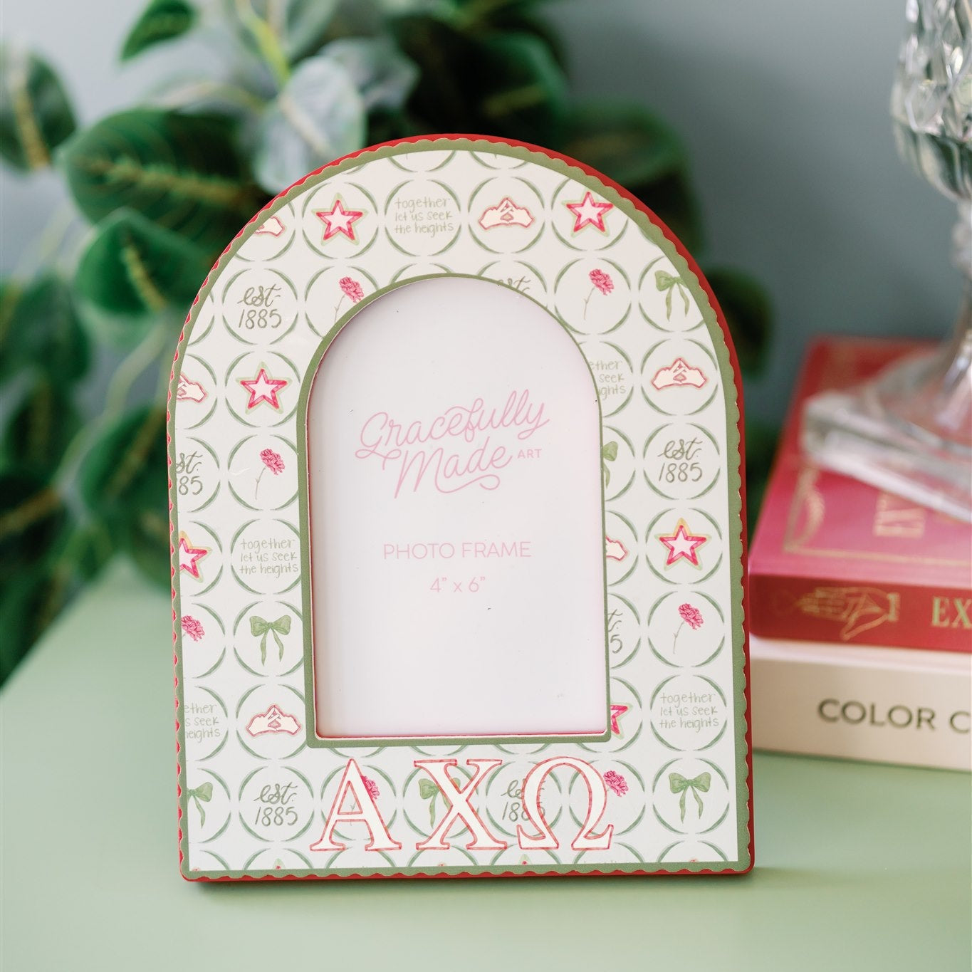 Decorative photo frame with 'Alpha Chi Omega' initials on a green surface with books and plants in the background.