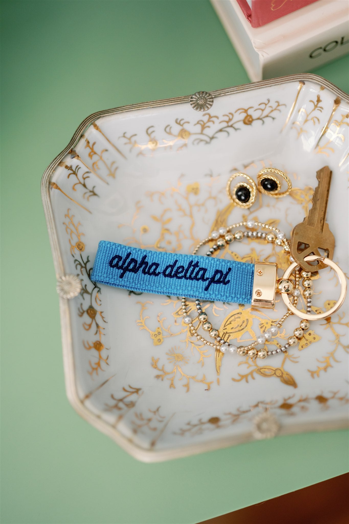 Decorative jewelry dish with gold jewelry and an Alpha Delta Pi blue keychain.