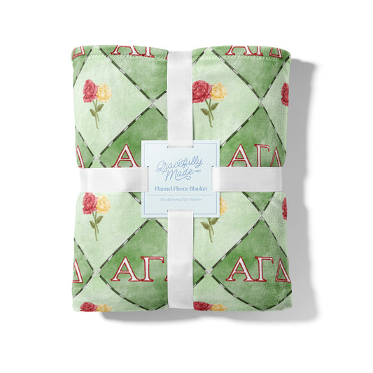 Folded green blanket with floral patterns and 'Alpha Gamma Delta' letters, tied with a white ribbon, on a white background.