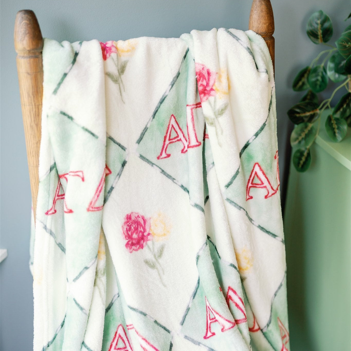 Floral patterned blanket with 'Alpha Gamma Delta' letters draped over a wooden chair against a gray wall.