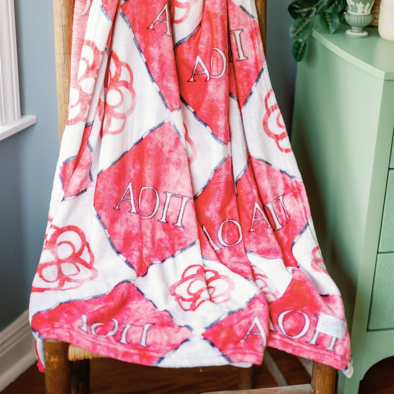 Red and white patterned blanket with Alpha Omicron Pi Greek letters draped over a wooden chair against a light blue wall.