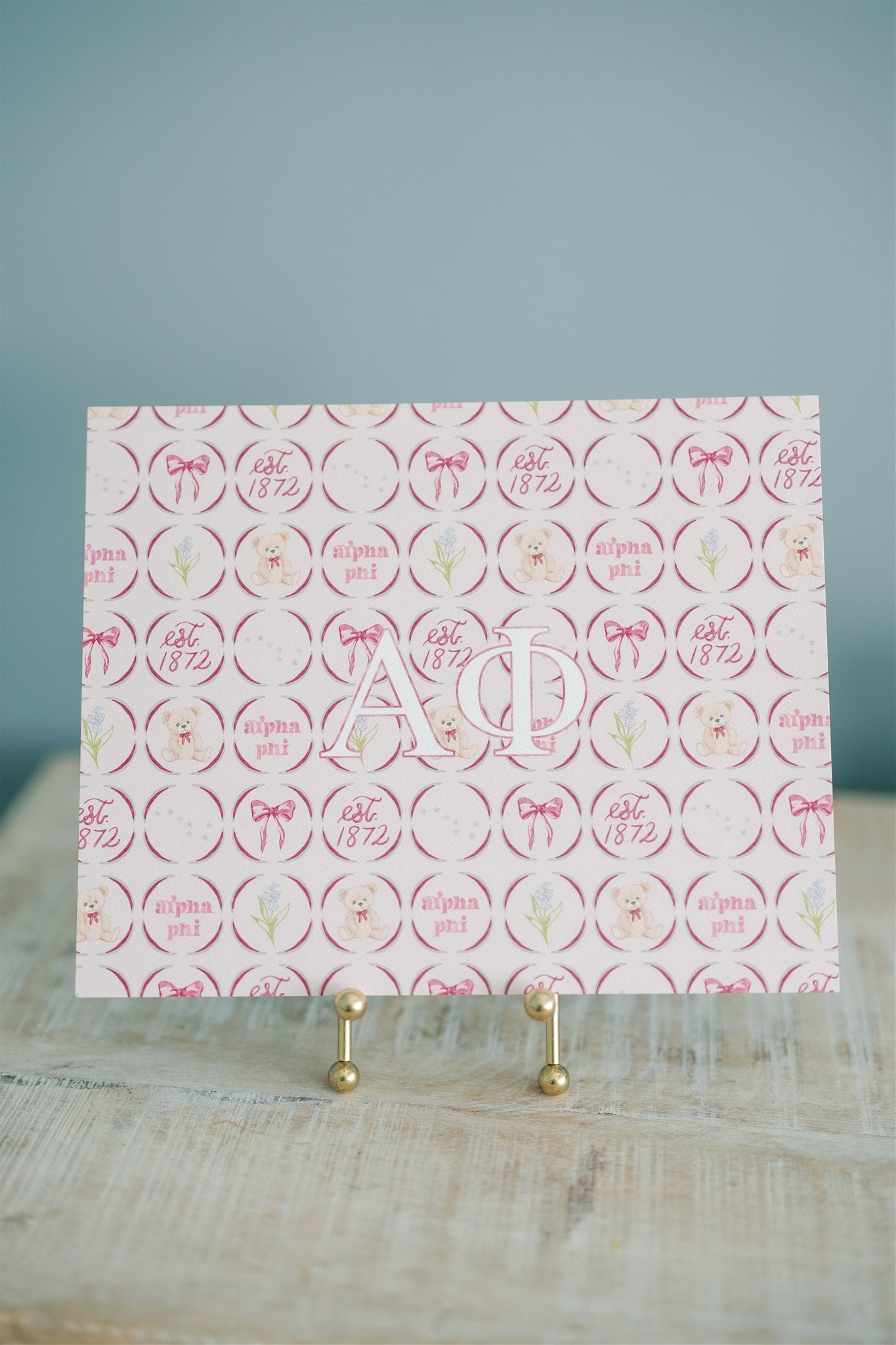 Decorative art print with 'Alpha Phi' Greek letters on a stand against a light blue background