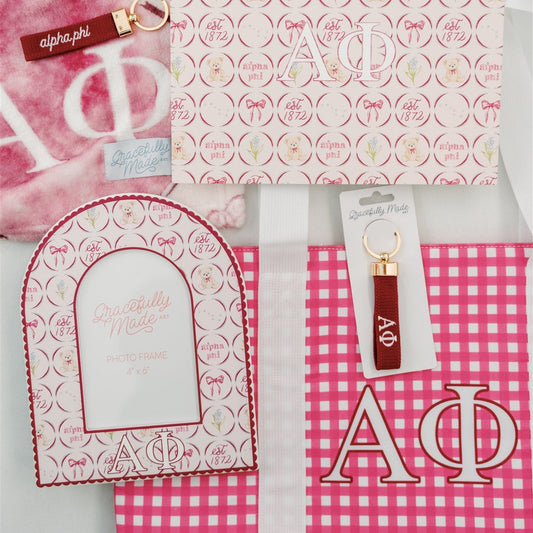 Collection of Alpha Phi merchandise including a blanket, keychain, and decorative items on a pink and white checkered background.