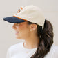 Woman wearing a beige and navy cap with a ponytail, against a plain background