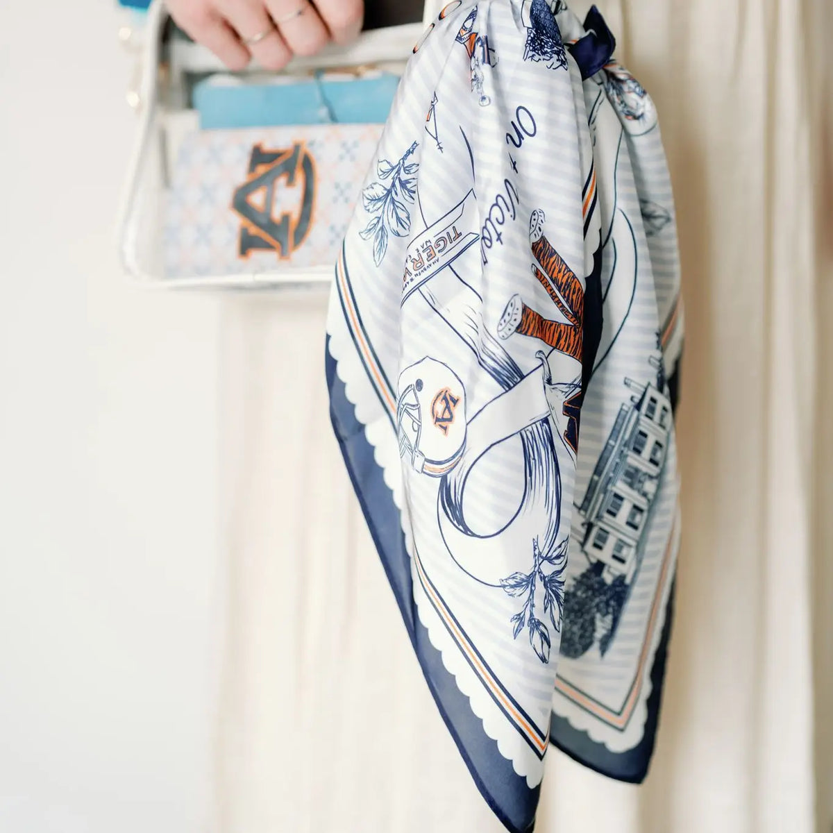 A close-up of a person holding a white silk scarf with a navy blue border and a light blue stripes print featuring Auburn University illustrations including a football helmet, toilet paper, and the tiger mascot.