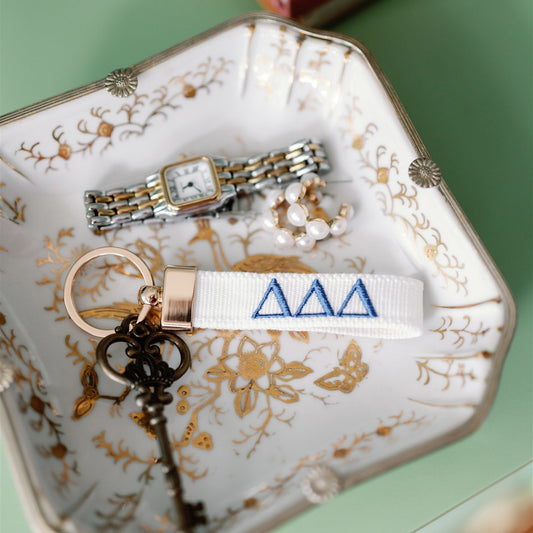 Decorative jewelry dish with jewelry and a white keychain with Tri Delta Greek letters embroidered on it