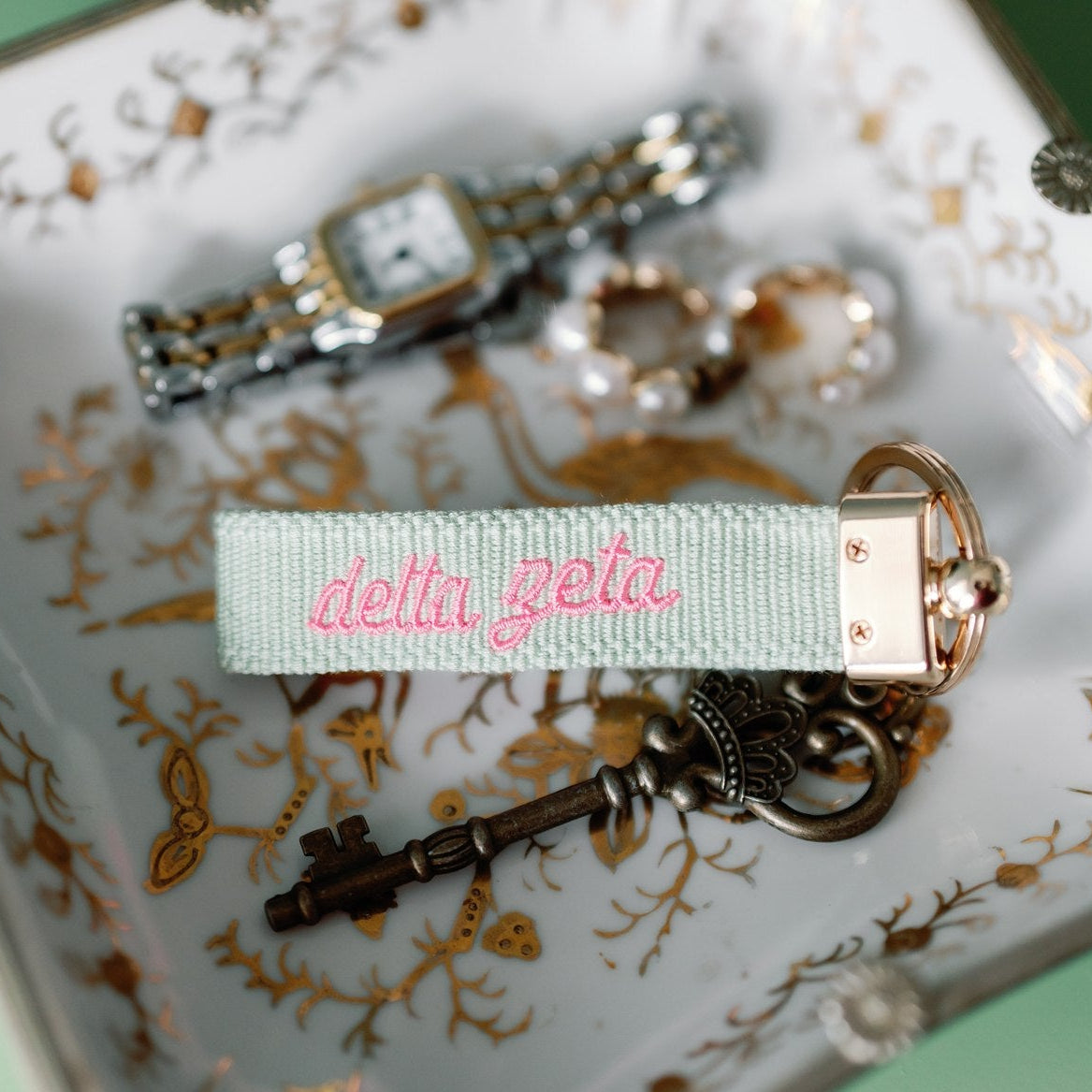 'delta zeta' embroidered in pink on a green keychain.ribbon on a decorative plate with jewelry