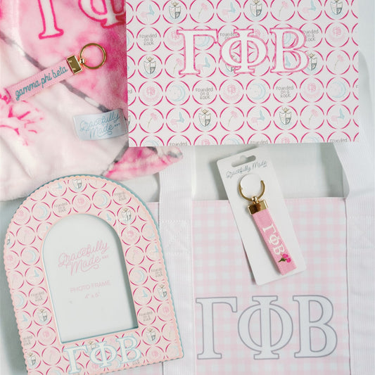 Gamma Phi Beta themed items including a pink keychain, tote bag, and decorative items on a matching pink and white patterned background.