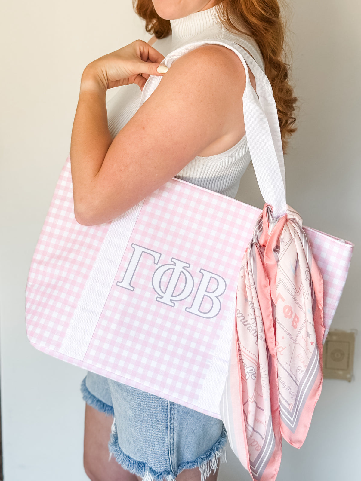 A woman wearing a white ribbed tank top and denim shorts carries a large pink and white gingham tote bag with white straps. The bag features the Greek letters Gamma Phi Beta (ΓΦB) in white on the front and has a matching pink and white printed scarf tied to the side.