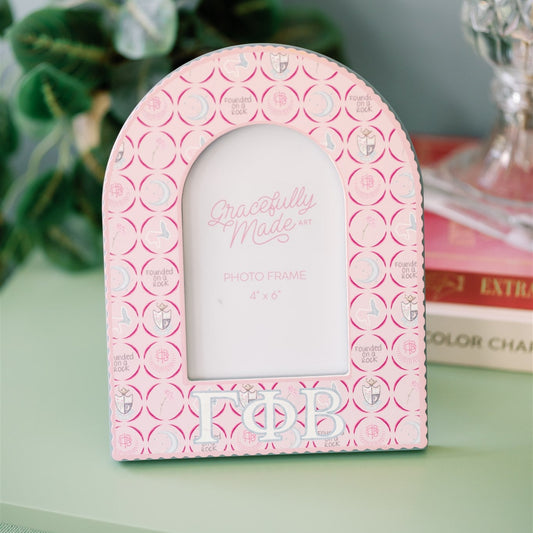 Pink decorative frame with Gamma Phi Beta pattern and Greek letters on a green surface.