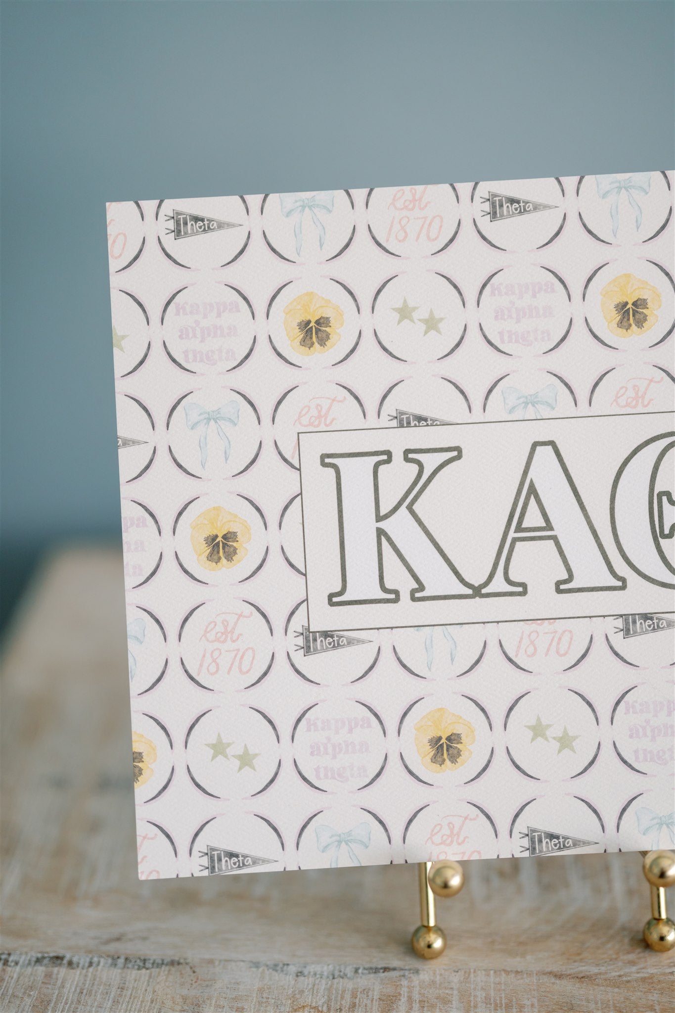 Decorative card with Kappa Alpha Theta Greek letters and circular designs on a stand against a blue background
