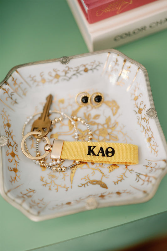 Decorative dish with keys, a bracelet, and a yellow keychain with Kappa Alpa Theta Greek letters embroidered on it