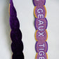 A close up of a beaded purse strap showing the front side with a purple background, scalloped border, and 'GEAUX TIGERS' text and the back of the strap featuring a felt backing displayed on a light grey background.