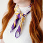 A close-up of a woman wearing a purple and gold striped silk scarf tied around her neck over a white ribbed top. The scarf features LSU themed illustrations and text.
