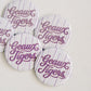 Five circle pin-back buttons with a white and light purple striped pattern, featuring the text 'Geaux Tigers' in purple cursive script, arranged on a light beige textured fabric background.