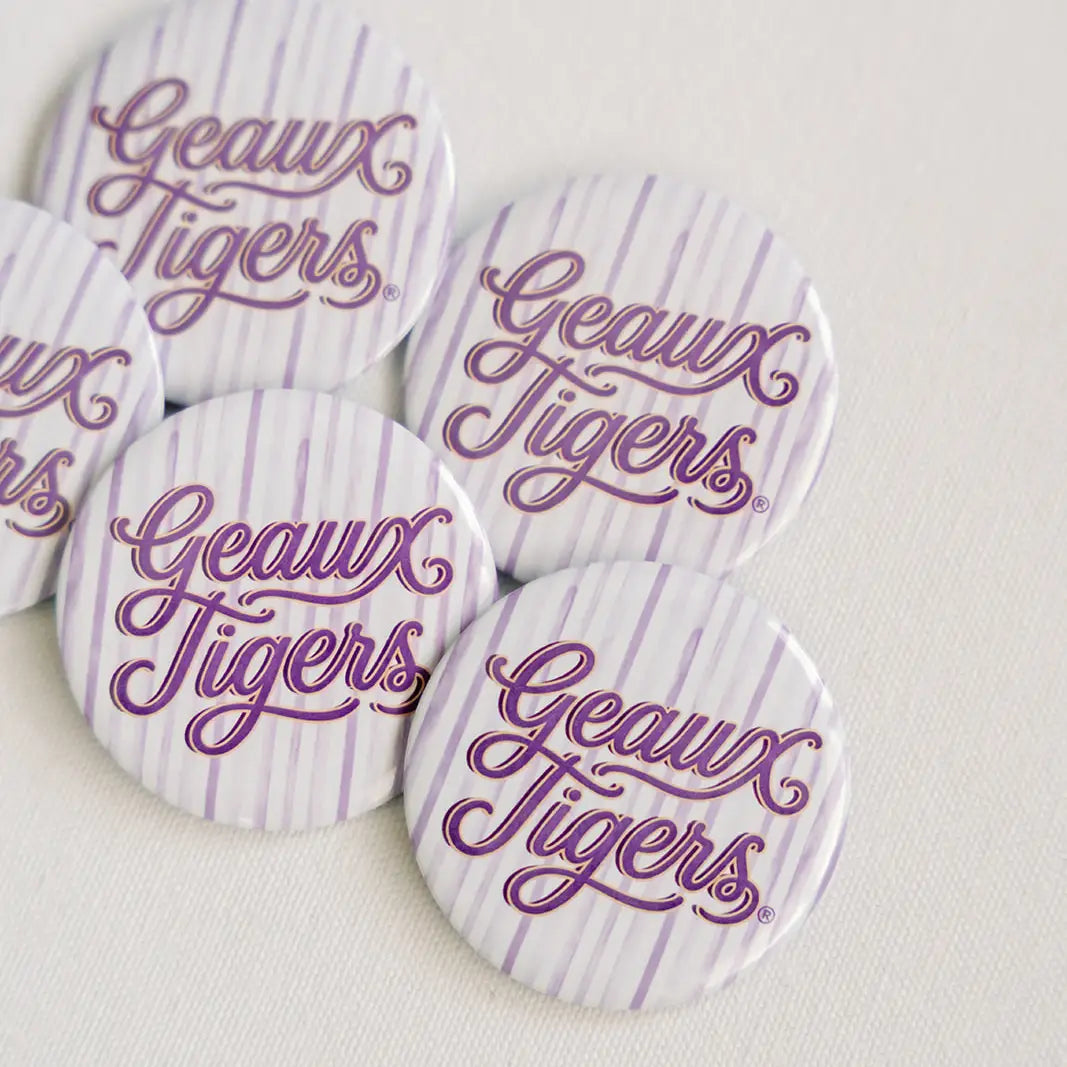 Five circle pin-back buttons with a white and light purple striped pattern, featuring the text 'Geaux Tigers' in purple cursive script, arranged on a light beige textured fabric background.