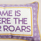 A close-up of a needlepoint pillow featuring the phrase 'HOME IS WHERE THE TIGER ROARS' in purple thread on a white background, framed by a purple patterned border and a thin yellow cord edge.