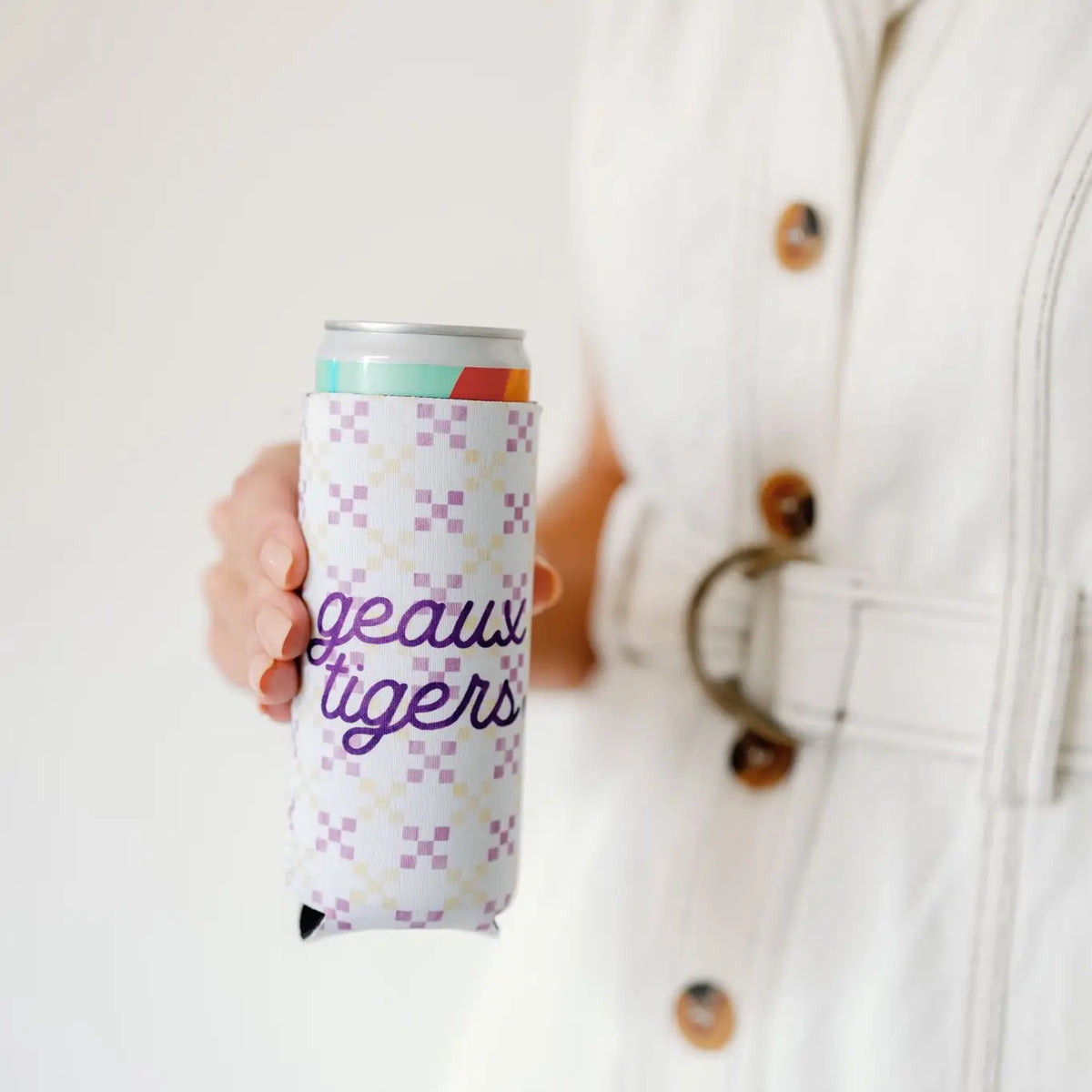 A person in a white belted coat holds a silver can inside a white can cooler with a purple and yellow checkerboard pattern and the words 'geaux tigers' in purple script.