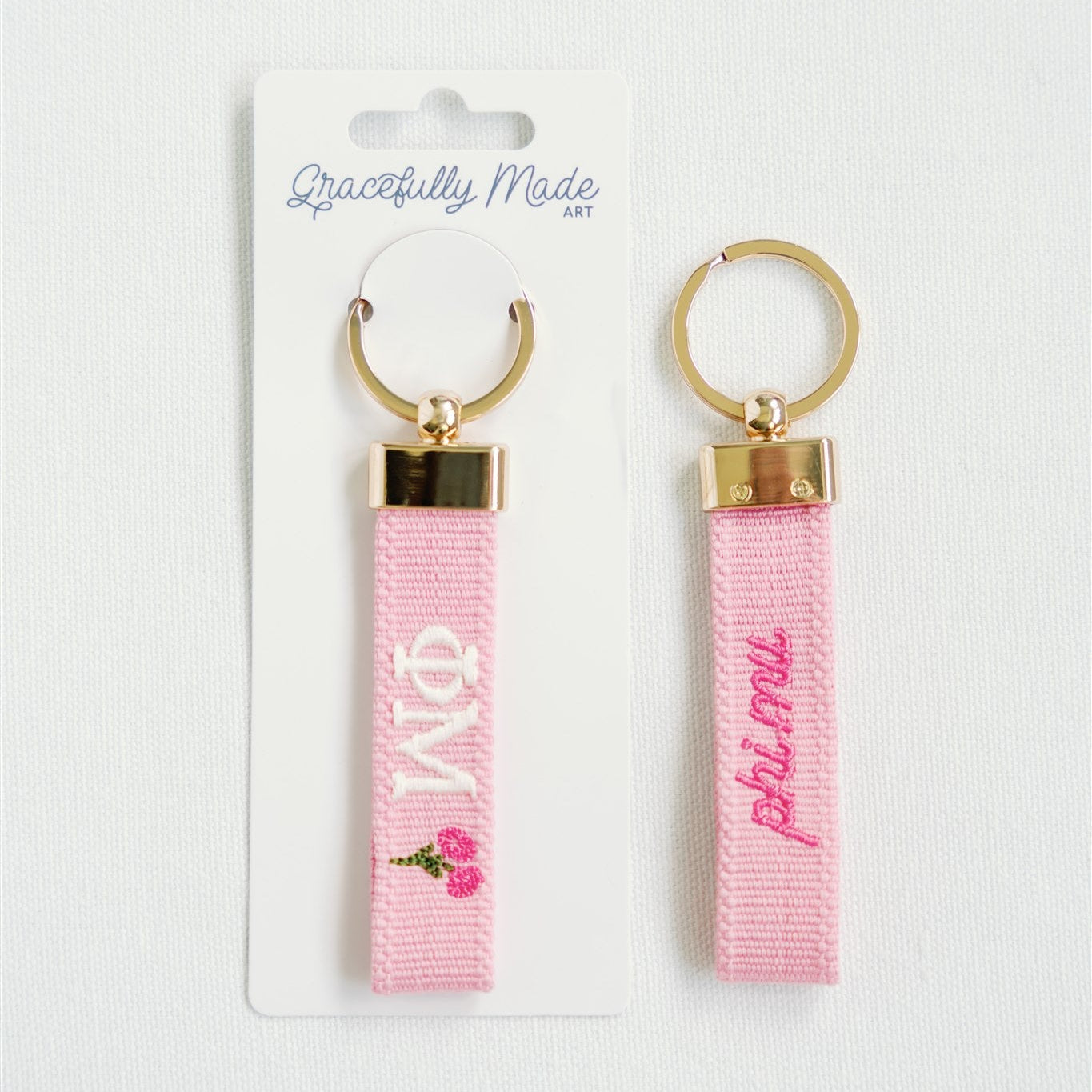 Front and back of a pink keychain with Phi Mu Greek letters and roses embroidered on it in Gracefully Made Art packaging.