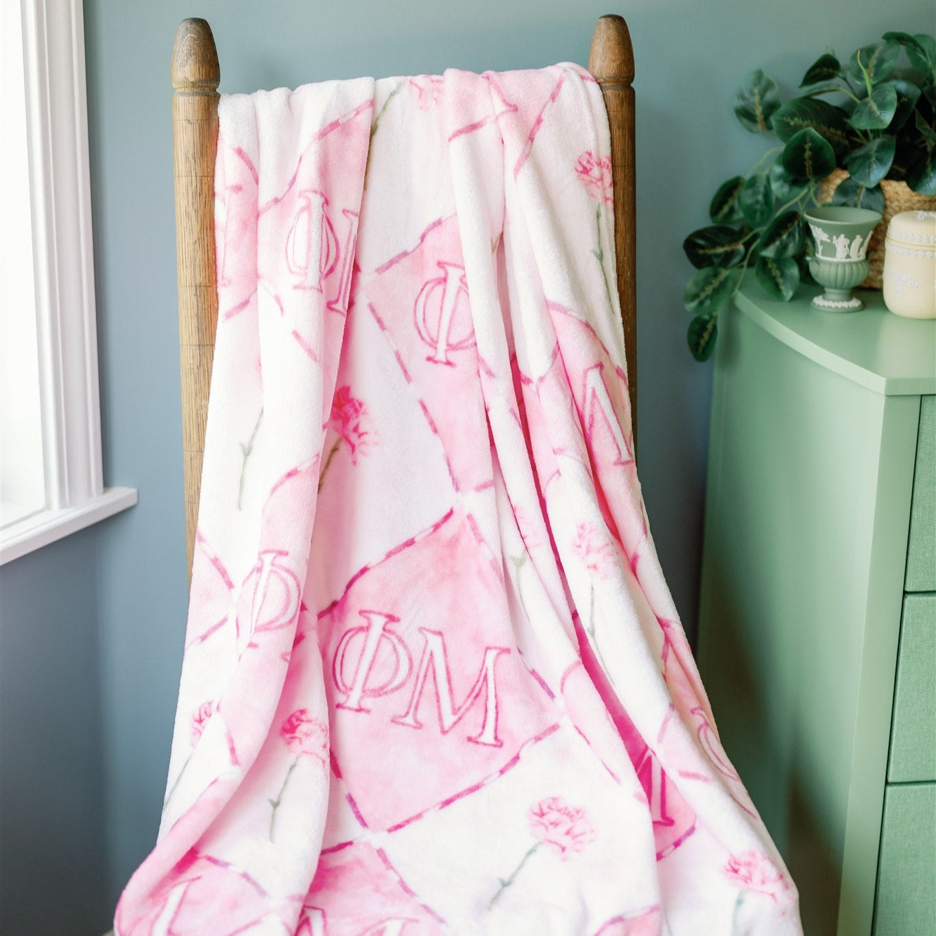 Pink and white Phi Mu patterned blanket draped over a wooden rack against a light blue wall.