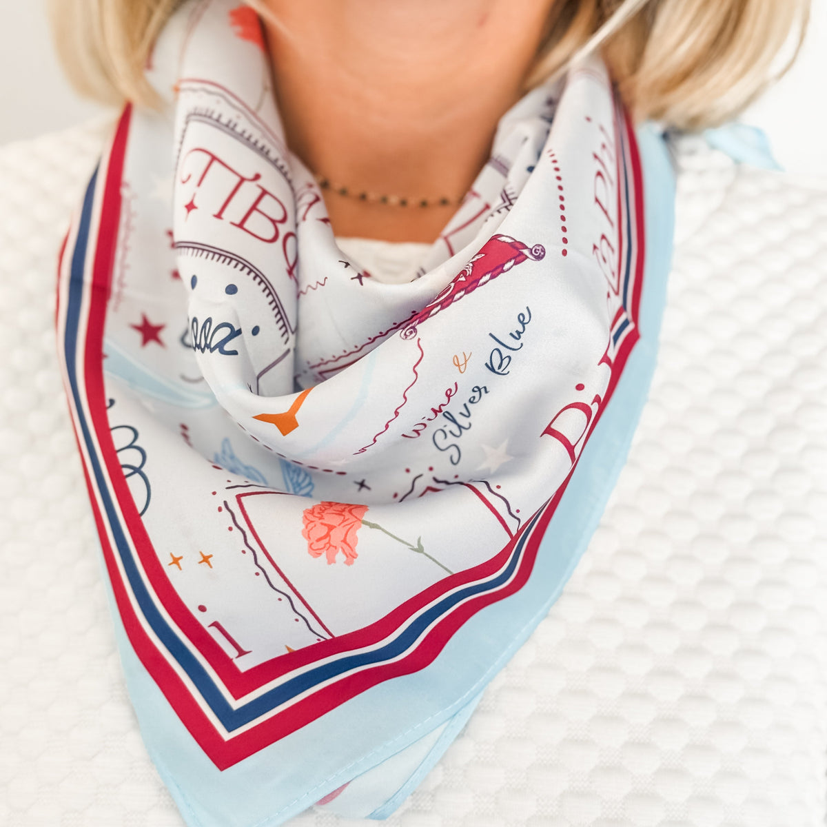 A close-up of a woman wearing Pi Beta Phi themed scarf with light blue, red, and navy text and illustrations, styled around her neck over a white textured top.