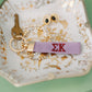 Purple keychain embroidered with Sigma Kappa Greek letters sitting on a jewelry dish.