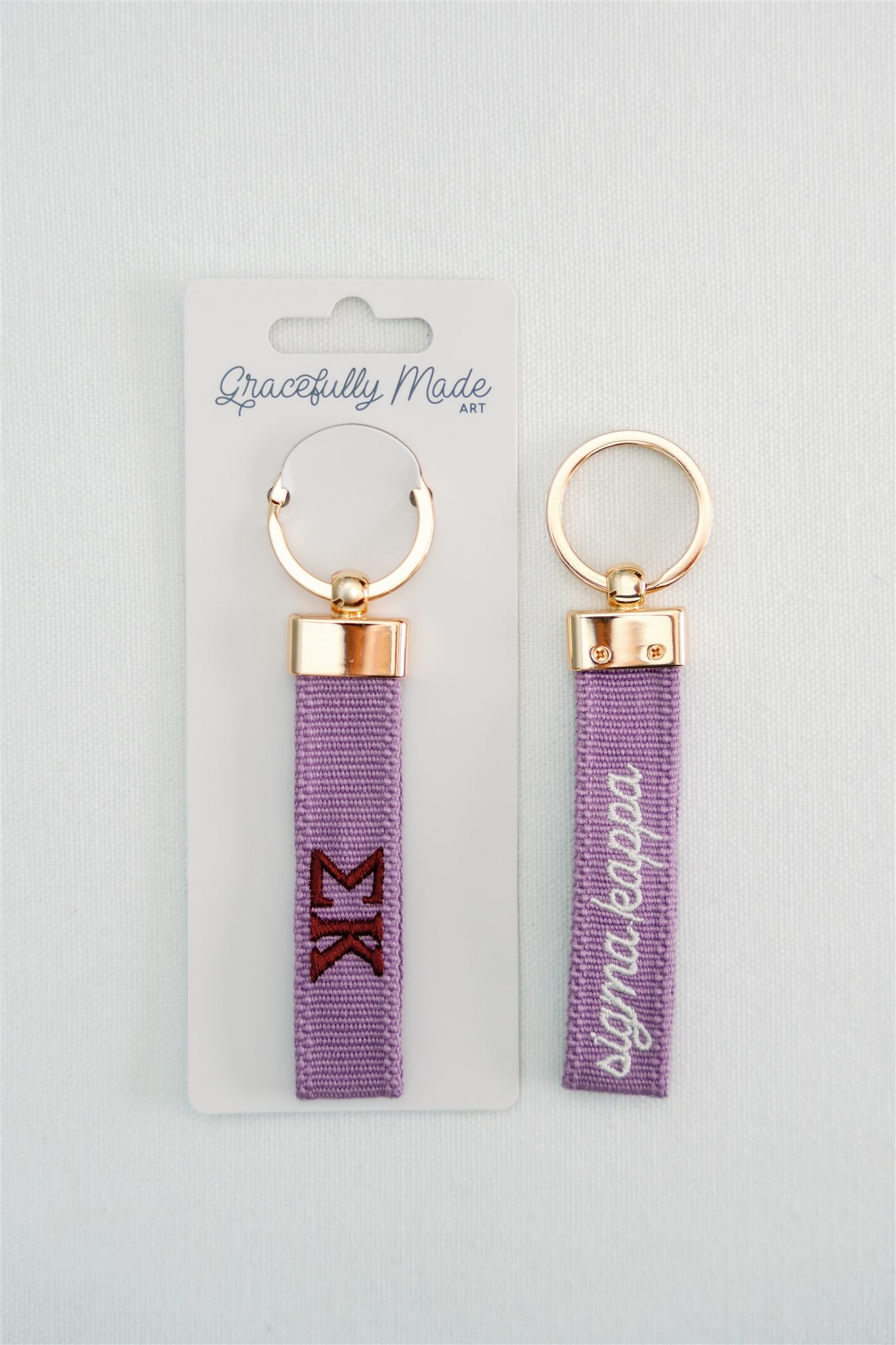 Front and back of a purple keychain with Sigma Kappa embroidered on it in Gracefully Made Art branded packaging. 