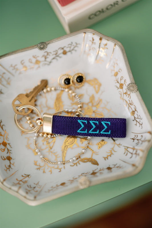 Blue keychain with Tri Sigma Greek letters embroidered on it set on a jewelry dish.