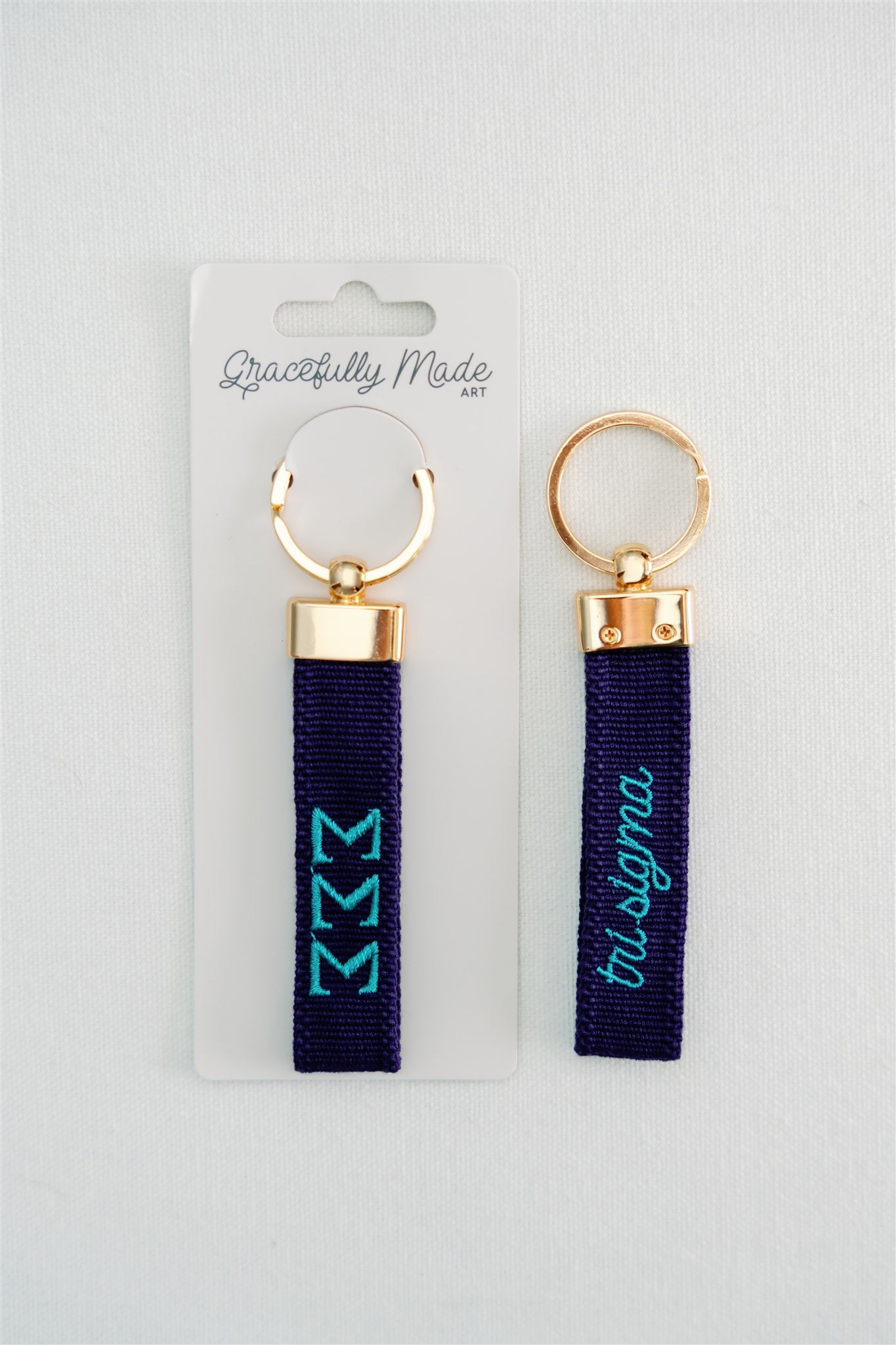 Front and back of a blue keychain with Tri Sigma embroidered on it in Gracefully Made Art packaging. 