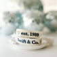 White woven bracelet with a gold bow and "Swift & Co" written in navy on one side and "est. 1989" written in navy on the other side.