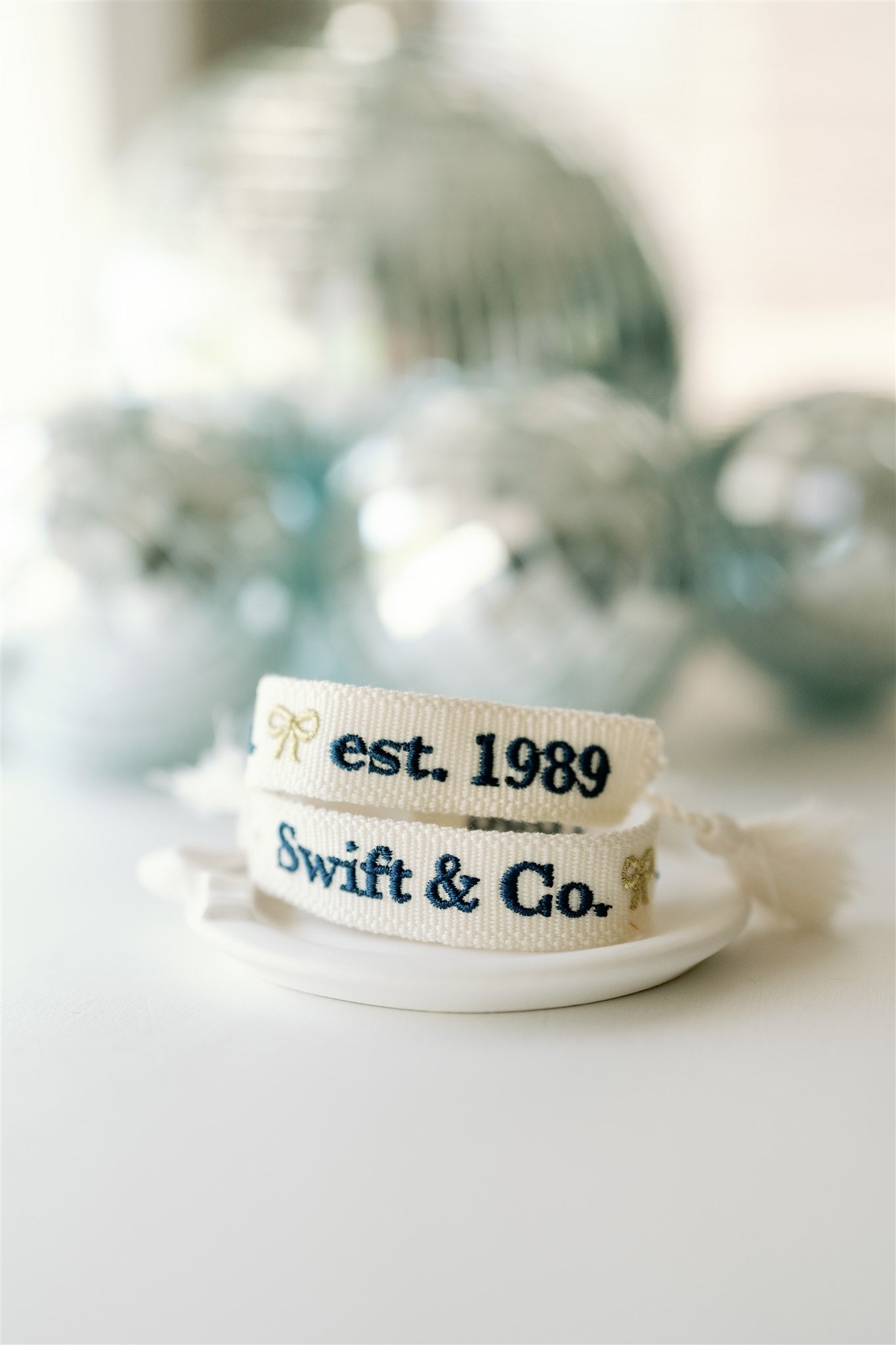 White woven bracelet with a gold bow and "Swift & Co" written in navy on one side and "est. 1989" written in navy on the other side.