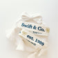 White woven bracelet with a gold bow and "Swift & Co" written in navy on one side and "est. 1989" written in navy on the other side.