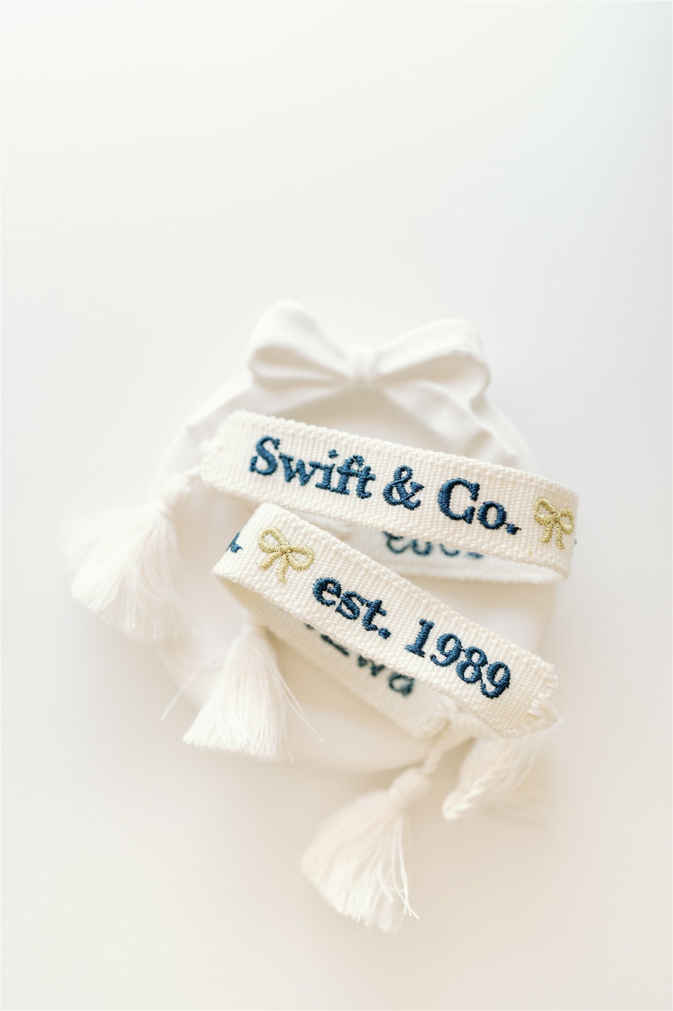White woven bracelet with a gold bow and "Swift & Co" written in navy on one side and "est. 1989" written in navy on the other side.