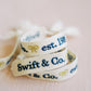 White woven bracelet with a gold bow and "Swift & Co" written in navy on one side and "est. 1989" written in navy on the other side.