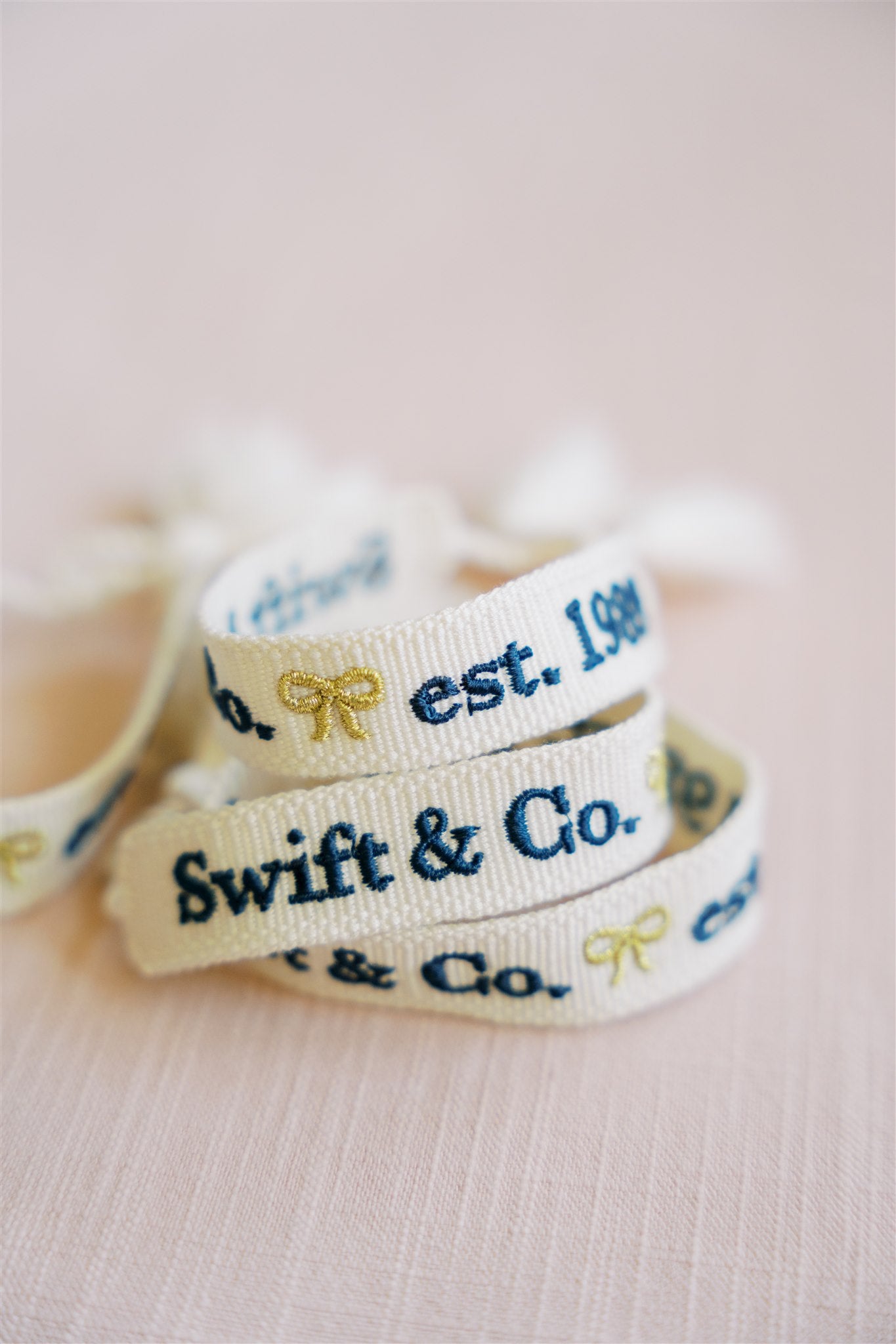 White woven bracelet with a gold bow and "Swift & Co" written in navy on one side and "est. 1989" written in navy on the other side.