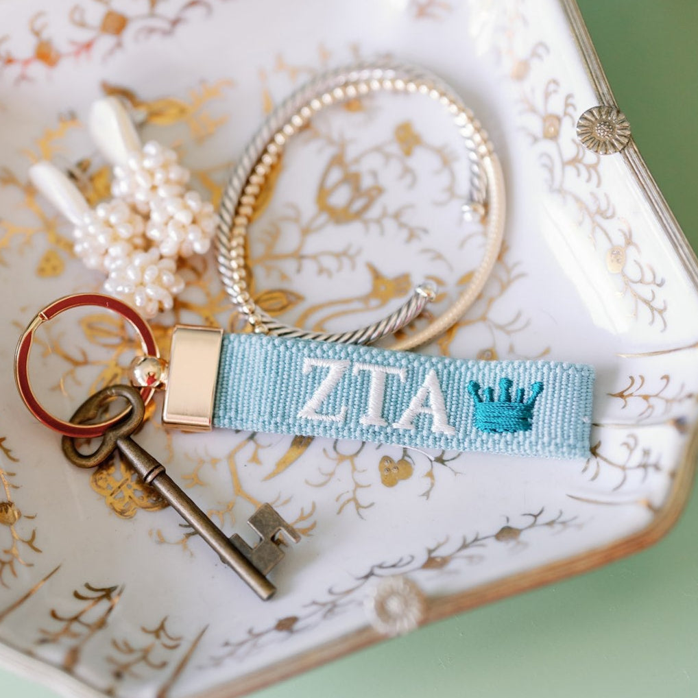 Light blue keychain with Zeta Greek letters embroidered on it set on a jewelry dish.