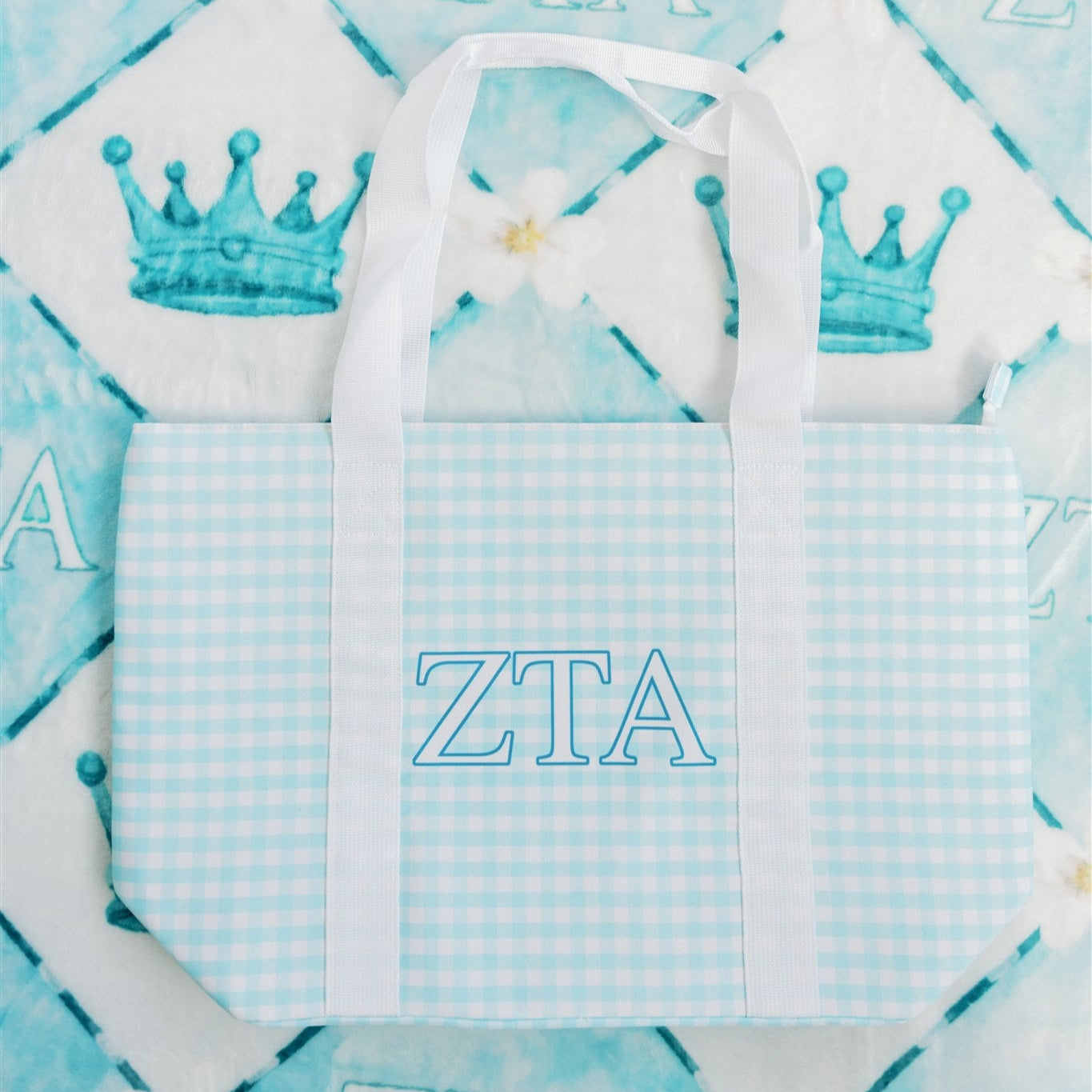 ZTA tote bag with blue gingham pattern, white handles, and Greek letters on a patterned background.